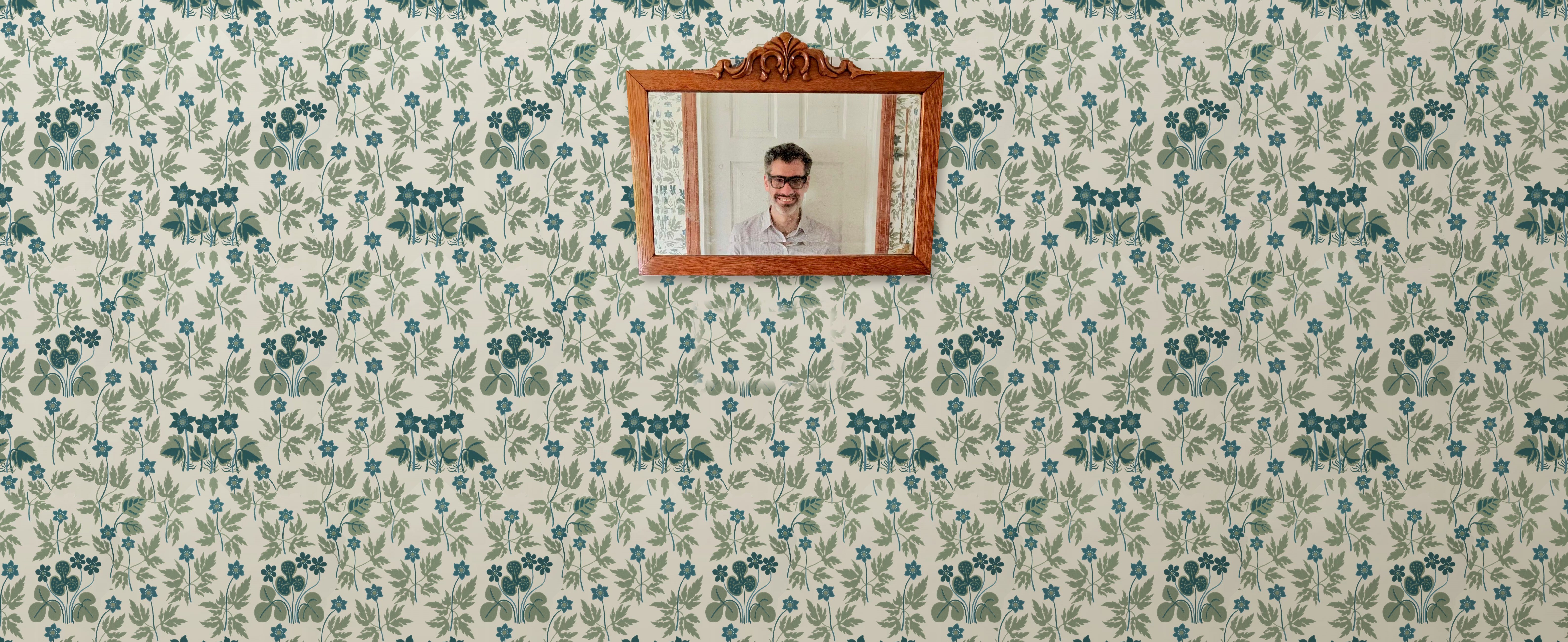 Hallway wall in blue-and-green floral wallpaper, an antique mirror reflecting Dan in glasses, and a thermostat reading 72°